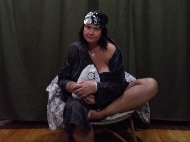 MiraaSweet with black hair, wearing a dark gray robe and black hat, sitting on a chair, holding a white pillow with a circular logo, showing her bare tits.