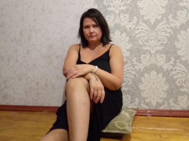 MiraaSweet with short black hair, wearing a black dress, sitting on a green pillow, arms folded, legs crossed, wooden floor, gray patterned wall, looking slightly annoyed.