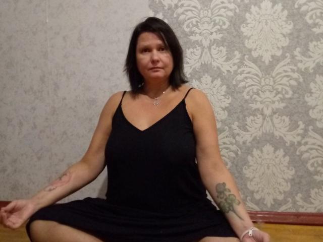 MiraaSweet, middle-aged woman with black shoulder-length hair, wearing a black dress, sitting with legs spread, tattoos on arms, neutral expression, wooden floor, patterned wallpaper in background.