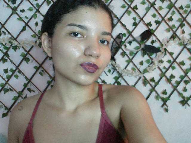 A selfie of SexLaddy with dark hair in a bun, wearing a red tank top, pouting with purple lipstick, showing a small tattoo on the right shoulder.