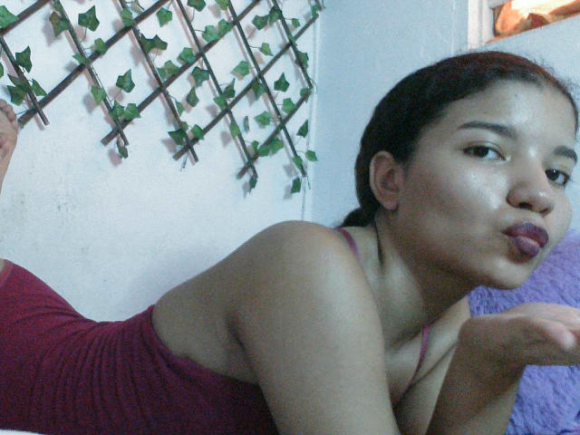 SexLaddy with dark hair in a braid, wearing a red dress, pouting with her lips, showing a bit of her ass, against a white wall with green vines.