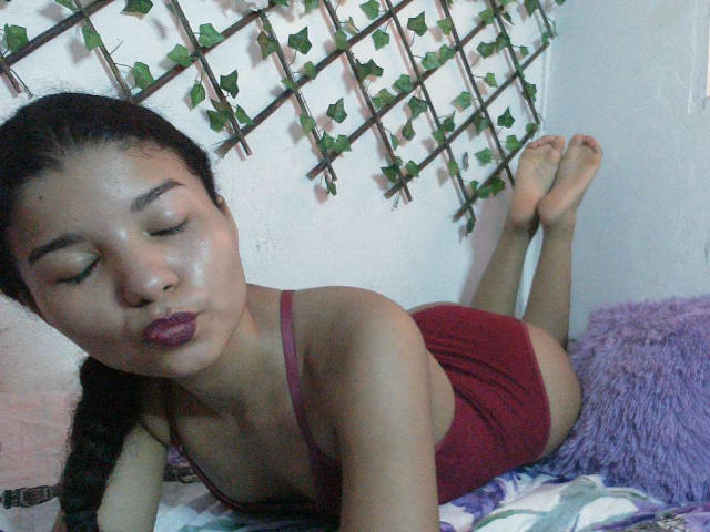 SexLaddy with closed eyes, purple lipstick, and red tank top, lying on purple pillow, butt up, legs crossed, black braid, soft skin, bare feet, wall with green vine pattern.