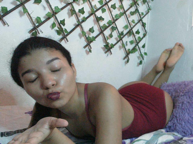 SexLaddy lying on her stomach, eyes closed, pouty lips, wearing a red tank top and shorts, showing her ass, with her hand in front, on a bed.