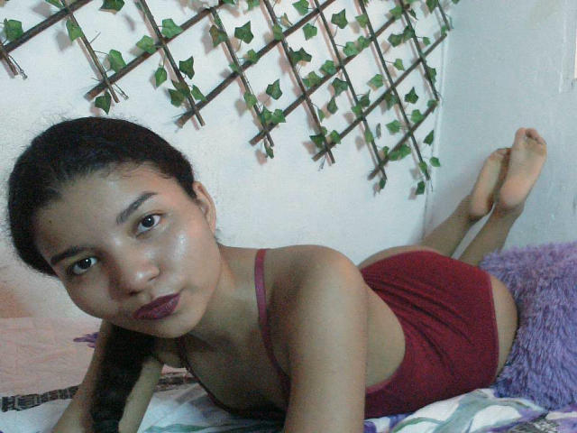 SexyLaddy lying on her stomach, wearing a red tank top and matching red booty shorts, with dark lipstick and a small nose. Her ass is up, feet in the air.