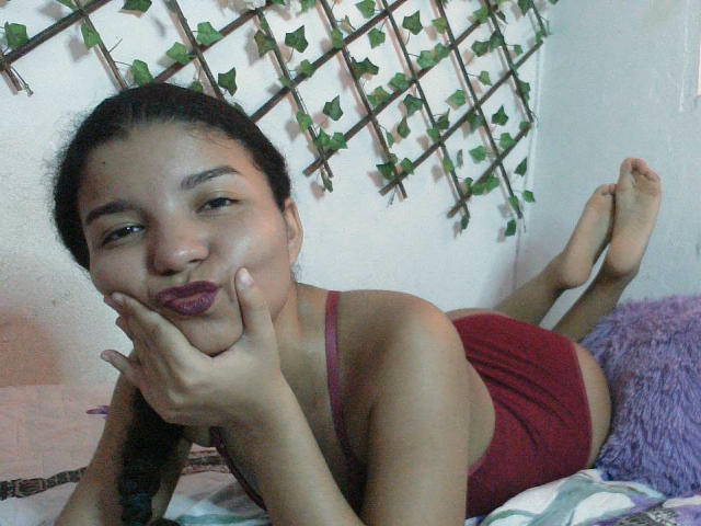 SexyLaddy with dark hair, lying on bed, wearing red underwear, purple lipstick, hand on cheek, legs up, looking at camera, against white wall with green vine pattern.