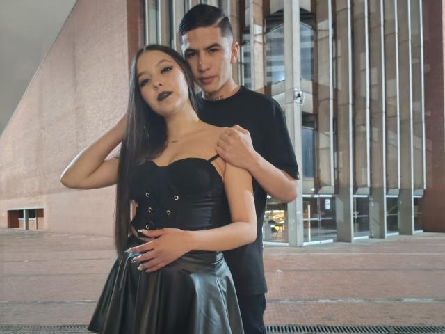 KylieAndJustin with long black hair, dark lipstick, black strapless leather dress, and Justin behind her, black t-shirt, hands on her shoulders, urban setting, Justin's short hair, Justin's eyes on her.