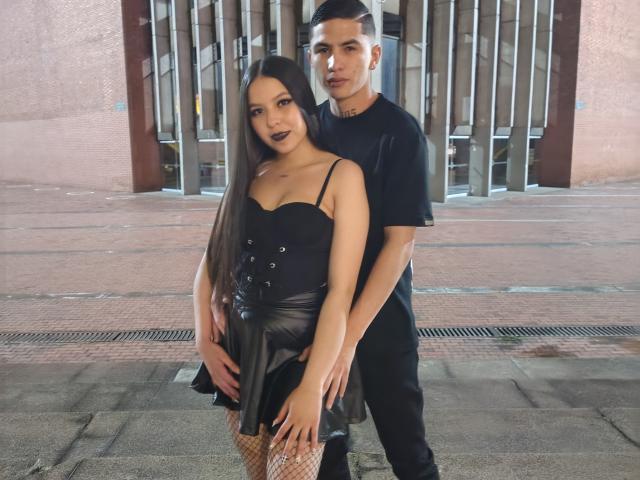 KylieAndJustin with long black hair, dark lipstick, black dress, and fishnet stockings, stands in front of Justin, who has short black hair, black shirt, and hands on her ass.