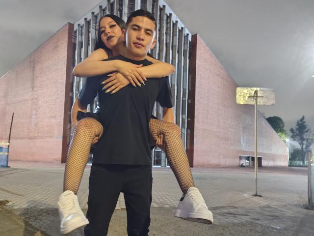 KylieAndJustin with tits in fishnet, black top, and white sneakers, riding Justin's shoulders, who's in black clothes, in front of a pink, brick building. Justin looks serious.