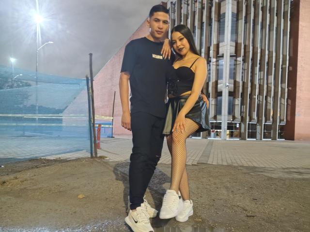 KylieAndJustin, with tits in a black top, ass in fishnet, and a green skirt, stands next to Justin, in black clothes, white sneakers, against a tall building.