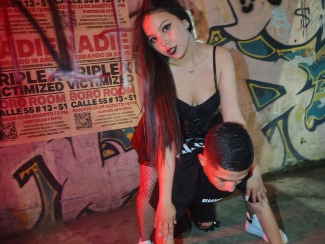 KylieAndJustin with long brown hair, black dress, and fishnet stockings, leaning over Justin's head, who has short black hair, wearing a black shirt, in a graffiti-laden room.