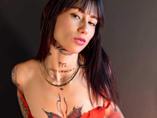 MiaNapoles, con cabello negro y lacio, flequillo y lápiz labial rosa, tiene tatuajes en el cuello y el pecho, usa una blusa de encaje rojo, un collar dorado y mira seductoramente hacia un lado..