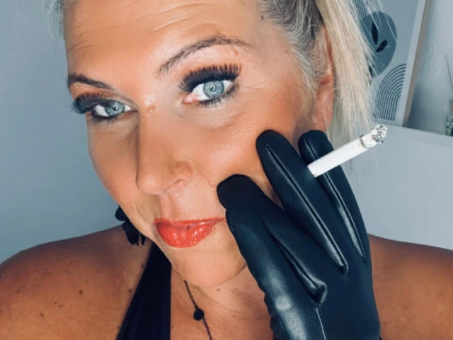 AurelieNoir, atractiva, con ojos azules, vestida con guantes de cuero negro, sosteniendo un cigarrillo encendido cerca de sus labios rojos y ahumados, mirando a la cámara con una ligera sonrisa..