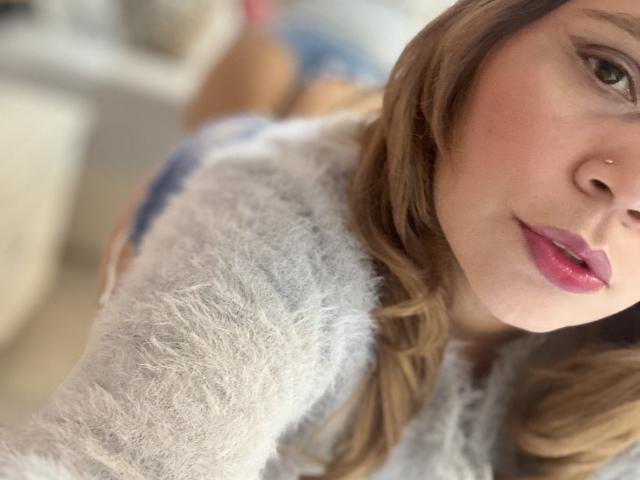 SophiaQueens with pink lips, nose piercing, and brown hair, wearing a fluffy white top, showing her tits slightly, in a blurry selfie. Her pink lipstick is glossy.