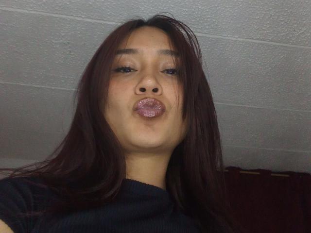 HanniaHot69 with long brown hair, puckering her shiny pink glitter lips, looking into the camera, wearing a black top, in a dimly lit room.