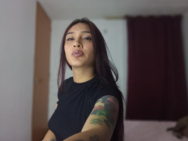 HanniaHot69 with long dark hair, pouty lips, black ribbed shirt, tattooed arm, and cute ass, standing in a room with a burgundy curtain.