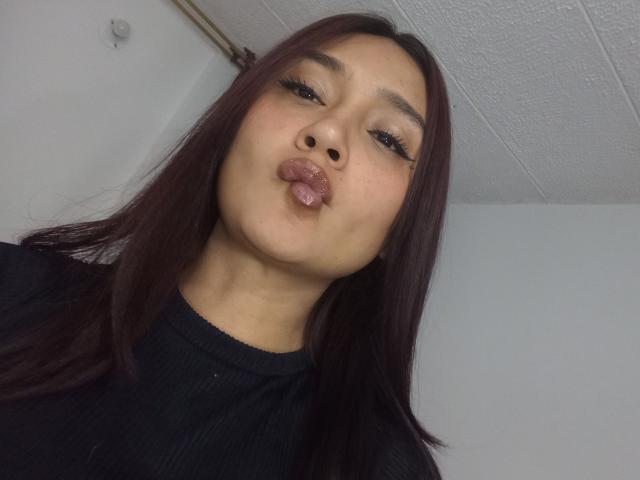 HanniaHot69 with dark brown hair, pouty lips, and long eyelashes, wearing a black shirt, making a pucker face. Her small nose and light skin are visible.