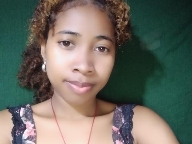 SexyEmilyStar69 with curly brown hair, light brown skin, wearing a lace tank top, small earrings, and a red necklace, smiling softly at the camera.