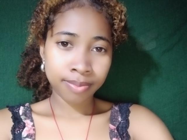 SexyEmilyStar69 has light brown skin, curly brown hair with blonde highlights, and is wearing a black lace tank top with pink floral patterns, looking at the camera.