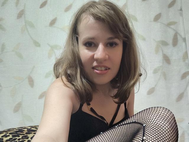 RosaJillian with medium brown hair, wearing black lace top and fishnet stockings, sitting with leopard-print cushion, showing small tits and slightly open mouth, looking at camera.