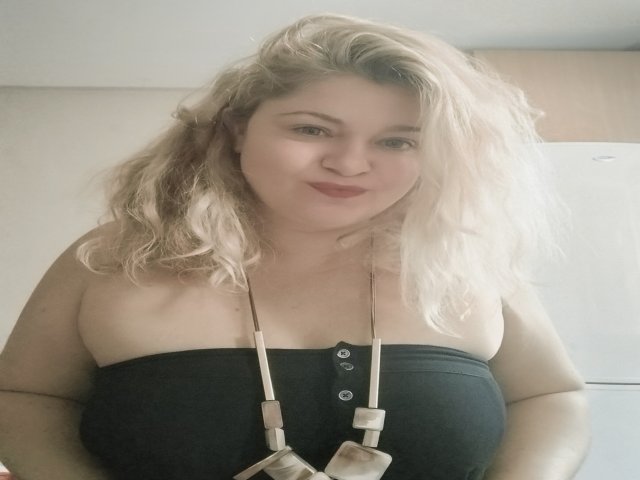 HurricaneCathrine has blonde, wavy hair, fair skin, and wears a black strap top showing her large boobs. She's smiling with red lipstick and a necklace in the photo.