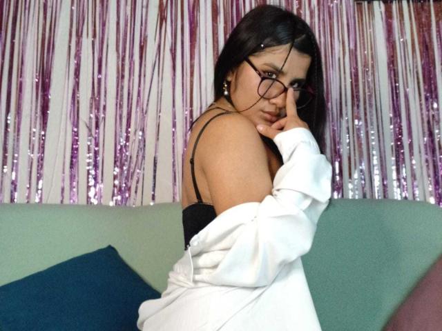 MelinoHottie with black hair, glasses, and a white shirt off her shoulders, showing her black bra and small, perky tits, looking over her shoulder with a slightly pouty face.