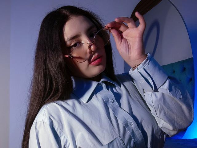 PaolaLyon with long dark hair, wearing glasses, adjusting them with her right hand, in a light blue button-up shirt, looking slightly to the side with pink lips.