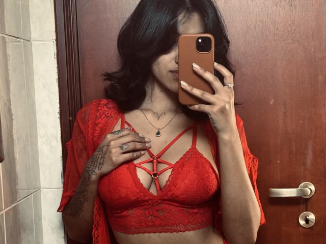 LollaCabral with black wavy hair, red lace bralette, strappy design, small heart tattoo on chest, holding phone, red lace shawl, tattoos on right hand, door in background.