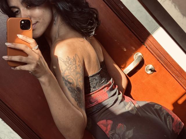 LollaCabral taking a mirror selfie in a tight, strapless, rose-patterned dress, showing a tattooed arm, holding an orange phone, with a ring on her left hand.