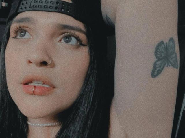 AgathaK with black hair, wearing a black cap, has a butterfly tattoo on her upper arm, nose ring, and choker, with slightly parted lips and a small lip piercing.