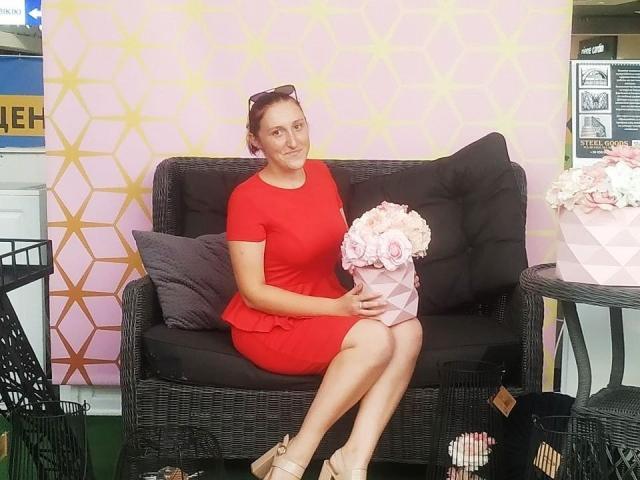 LiliTrayya in a red dress, sitting on a black wicker couch, holding pink and white roses, with sunglasses on her head, wearing beige wedge sandals, smiling.