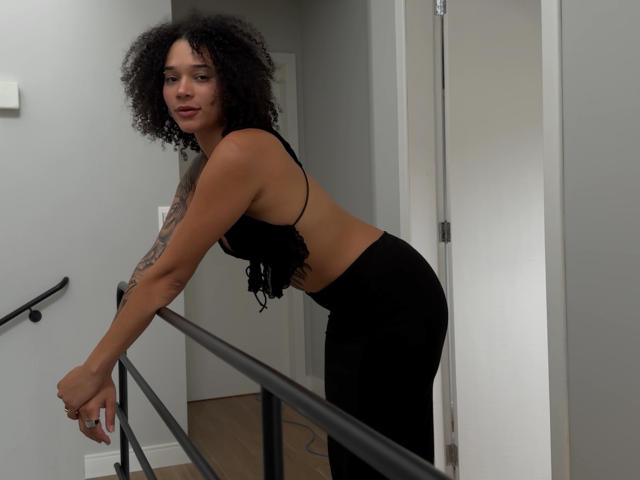 MauiMoor with curly black hair, tan skin, tattoos on right arm, wearing a black crop top and black pants, leaning on a black railing, looking at the camera.