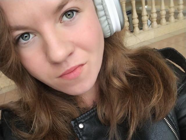 PrettyAhVoron has light green eyes, a slight smile, and is wearing white headphones with brown wavy hair, a black leather jacket, and no visible tits or ass.
