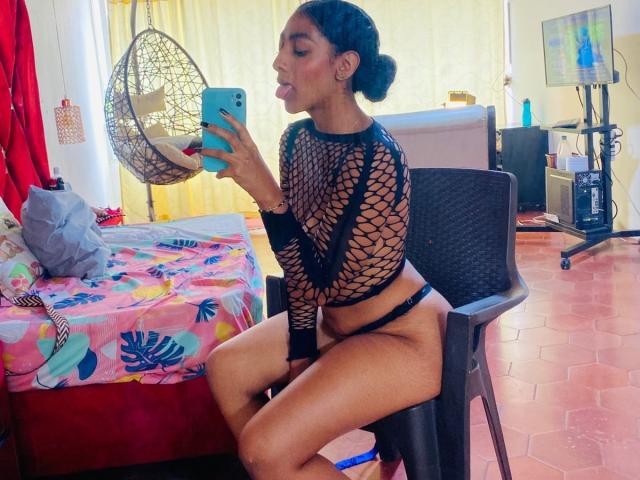 NatalyBlonder with dark hair in a bun, wearing a black fishnet top and black thong, sitting on a chair, taking a selfie with her tongue out, showing her ass.