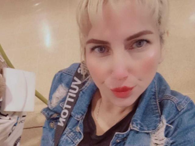 Blond GiuliaAddams with short hair, red lipstick, wearing a blue denim jacket, black shirt, and strap with "NOTION" on it, looking into camera.