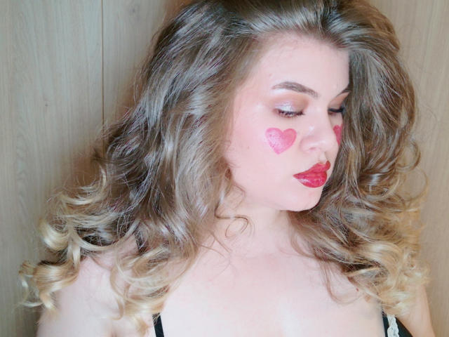 SweetnessX69 avec des cheveux blonds ondulés, un maquillage en forme de cœur rose sur les joues, du rouge à lèvres rouge, les yeux fermés, portant un bandeau noir, fond en bois clair, regardant vers le bas, lumière douce..