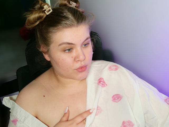 SweetnessX69 with light skin, blonde hair in double buns, wearing a white robe with pink lipstick prints, looking sideways, slightly pouting, hand on chest, nails long.