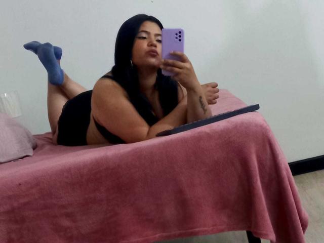 EmilyXGrace, with dark hair, lying on a pink bed, taking a mirror selfie, wearing a black top, blue socks, and holding a laptop, showing her ass and tattoos.