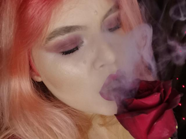 SweetnessX69 with pink hair, smoky eyeshadow, and white makeup, blows smoke into a red rose, her eyes closed, looking sexy and seductive.