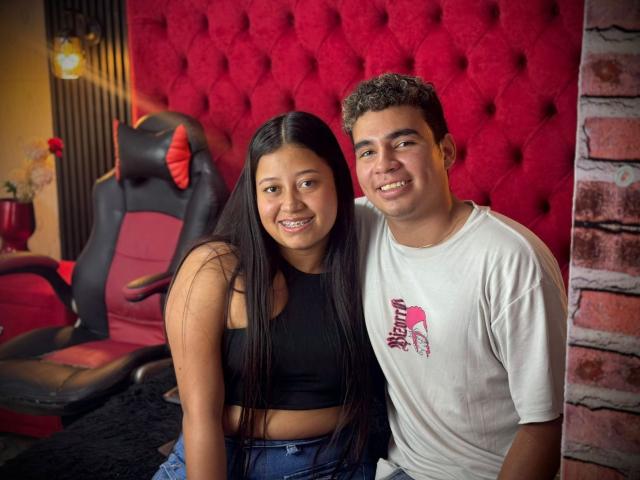 NatyAndYerry, with long black hair and small tits, sits next to Yerry, in a white tee, on a black bed, smiling. Yerry's hand is on NatyAndYerry's ass.