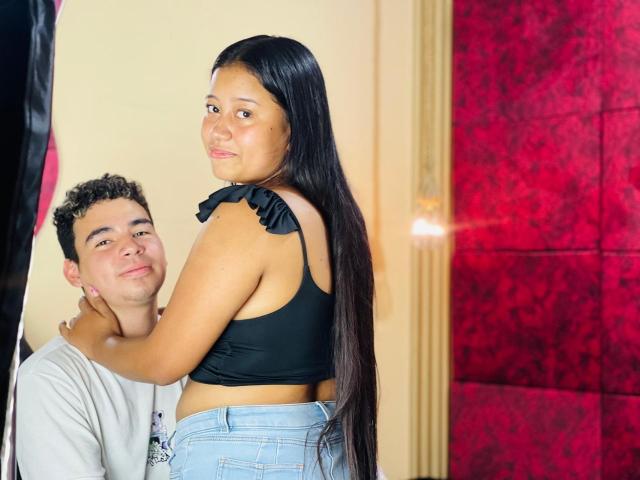 Naty has long black hair, wearing a black crop top with ruffled shoulder and blue jeans. Yerry, curly-haired, in a gray shirt, holds her by the neck, smiling.