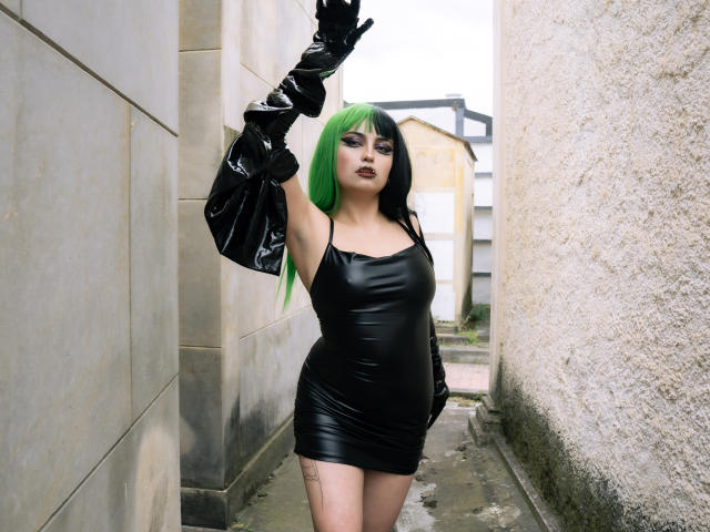 AngelBlakee with green and black hair, wearing a tight black latex dress and long black latex gloves, standing in a narrow alley, arms raised, showing her tits and ass.
