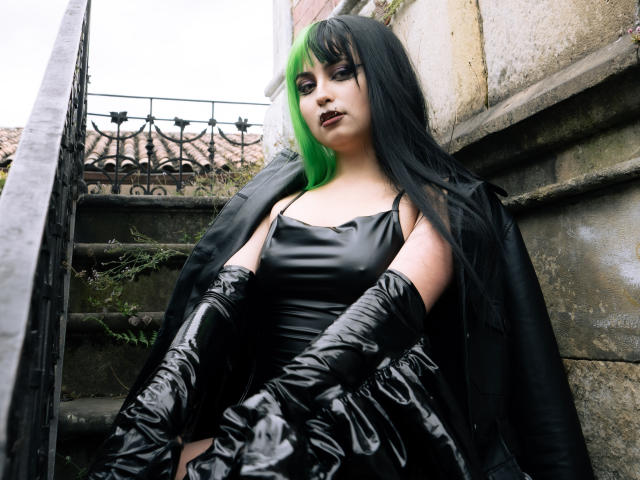 AngelBlakee with green and black hair, black leather dress, and gloves, sitting on stone steps, looking up with dark lipstick and smoky eye makeup. Her tits are visible.