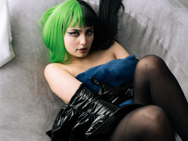 AngelBlakee with green and black hair, black eyeliner, blue tube top, black shiny gloves, and black pantyhose, sitting on a gray couch, looking up with a slightly open mouth.