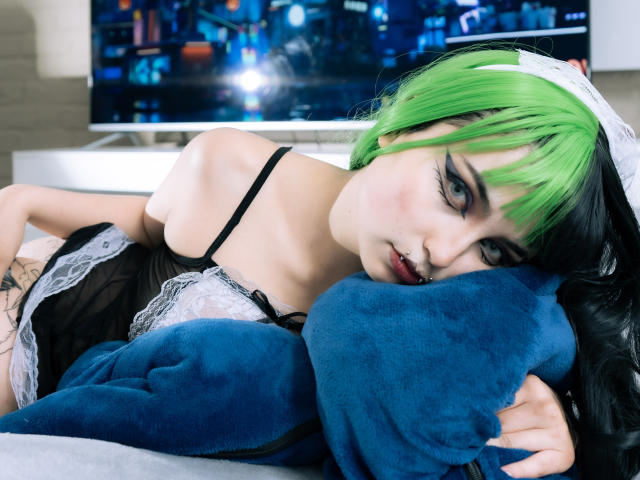 AngelBlakee with green and black hair, lying on a blue pillow, wearing a black lace maid outfit, black eyeliner, and a nose ring, looking seductively at the camera.