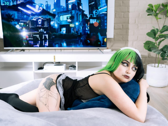 AngelBlakee with green and black hair, black tank top, and white lace panties, laying on bed, butt tattoo, black knee socks, TV in background showing neon city.