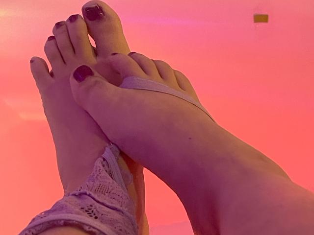 SamantaMuller's toes with dark purple nail polish, crossed at the ankles, wearing a lace garter, under pink lighting.