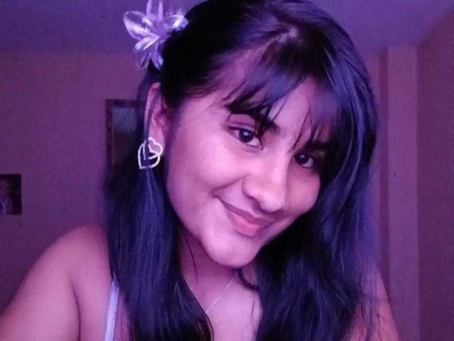 MelinoHottie with dark blue hair, purple ribbon, heart earrings, smiling at camera, wearing white tank top, purple lighting, cute face, slightly messy hair, soft smile, attractive.