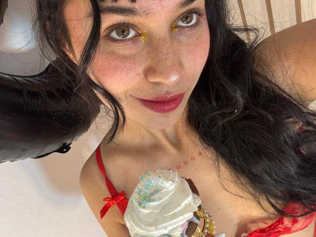 SolOrtega, avec ses cheveux foncés, ses taches de rousseur et son maquillage doré pour les yeux, tenant une glace au chocolat avec des vermicelles, portant de la lingerie rouge et souriant à l'appareil photo. Seins visibles..
