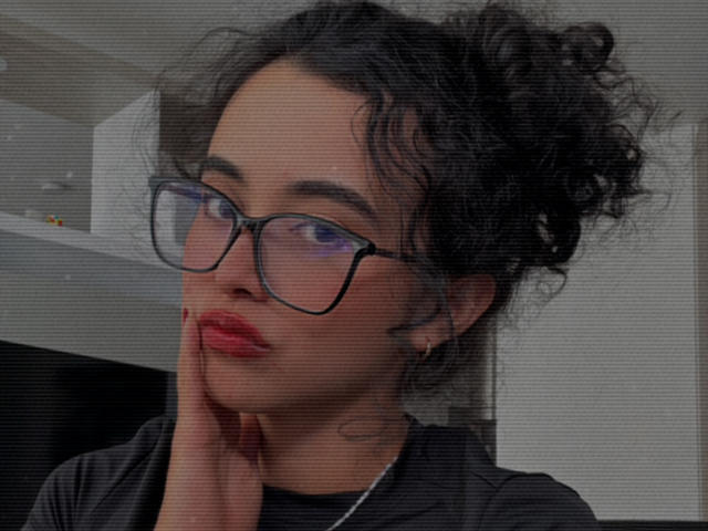 Vivienne Laurent heeft krullend zwart haar, draagt een zwarte bril, rode lippenstift en een zwart shirt, met haar hand in de buurt van haar kin, en kijkt iets opzij..