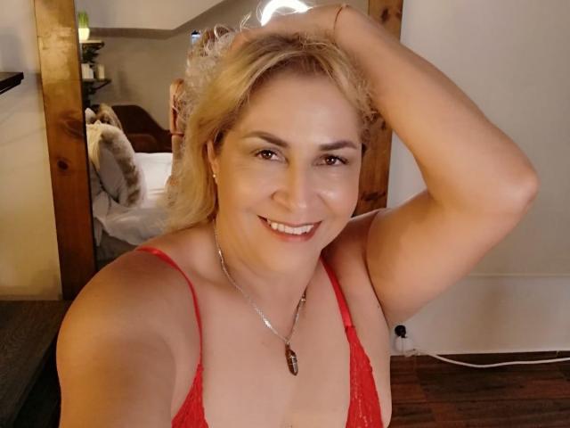 MajaReal with blonde hair, smiling, wearing a red lace bra, holding her hair up, showing her tits, and a necklace with a brown pendant, in a cozy room.